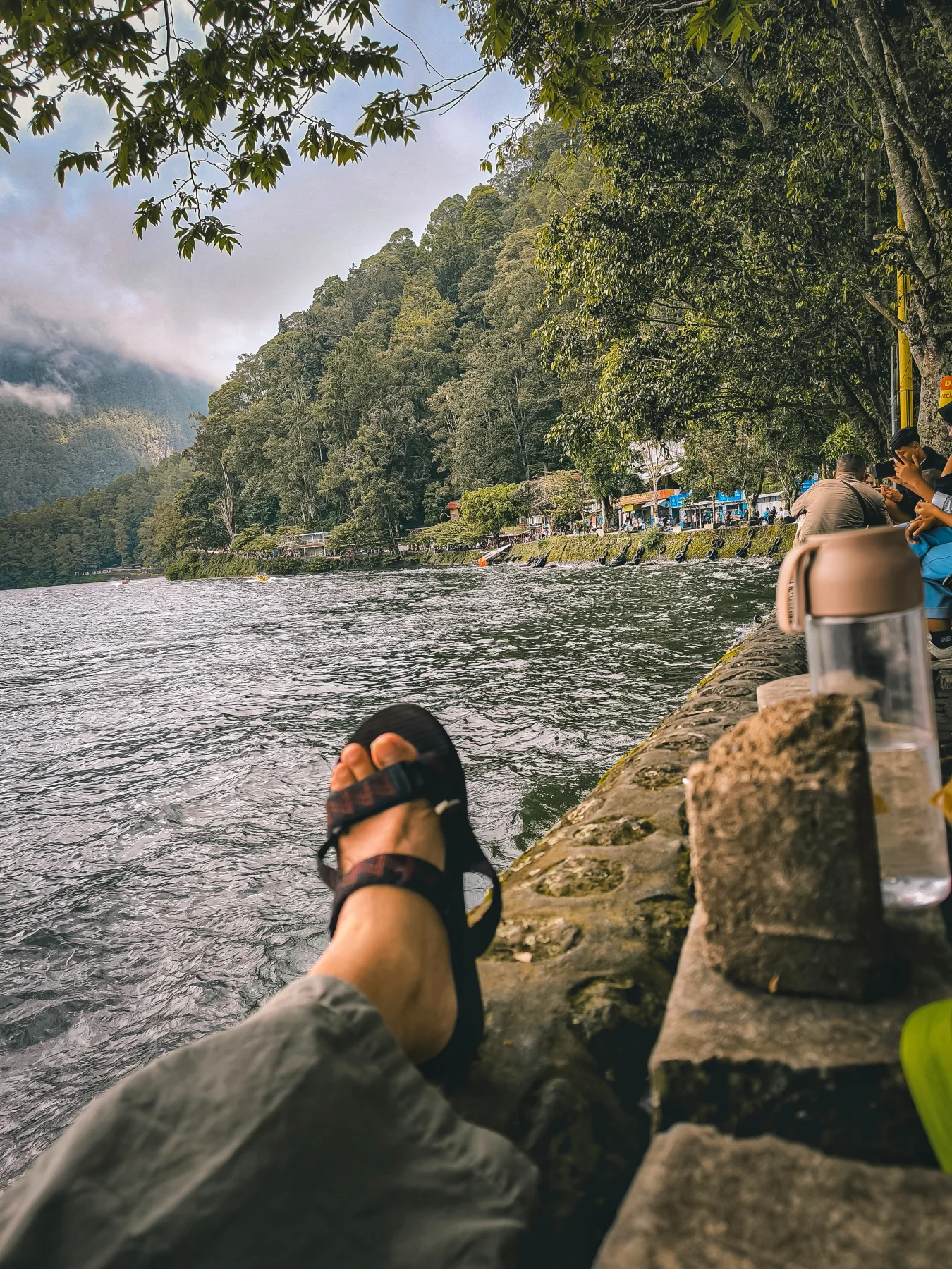 Pemandangan Telaga Sarangan dengan kaki bersandal di tepi telaga dan bukit hijau di seberang